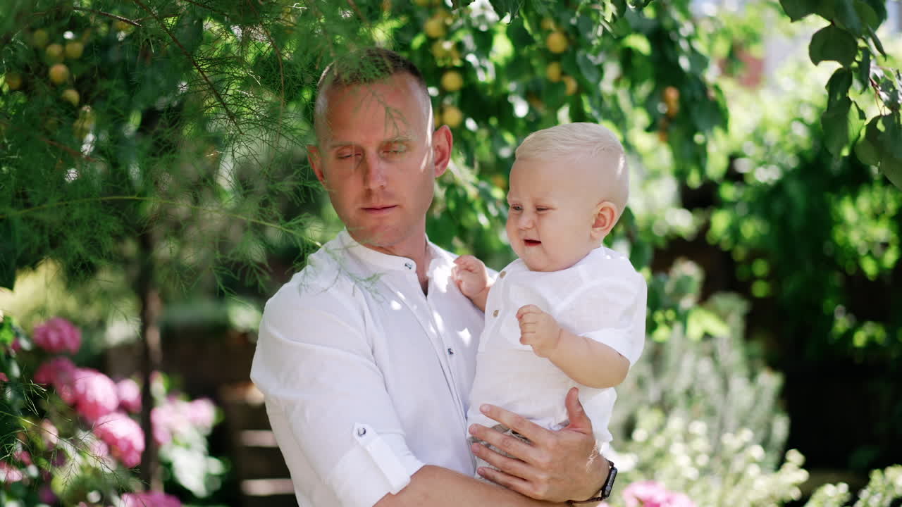 Caucasian daddy is holding his lovely baby outdoors. Father shows the tree to his child and kid touches the branch.