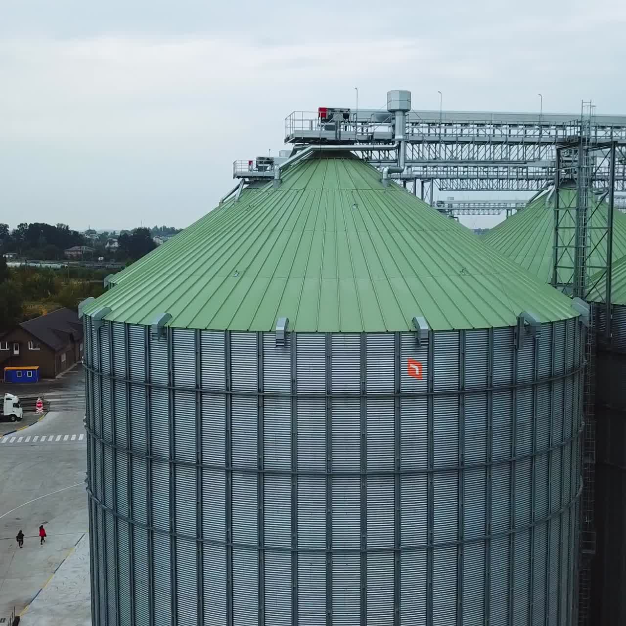 Metal tanks of elevator with diverse agricultural machinery nearby. People walking along the huge silver cisterns. View from above