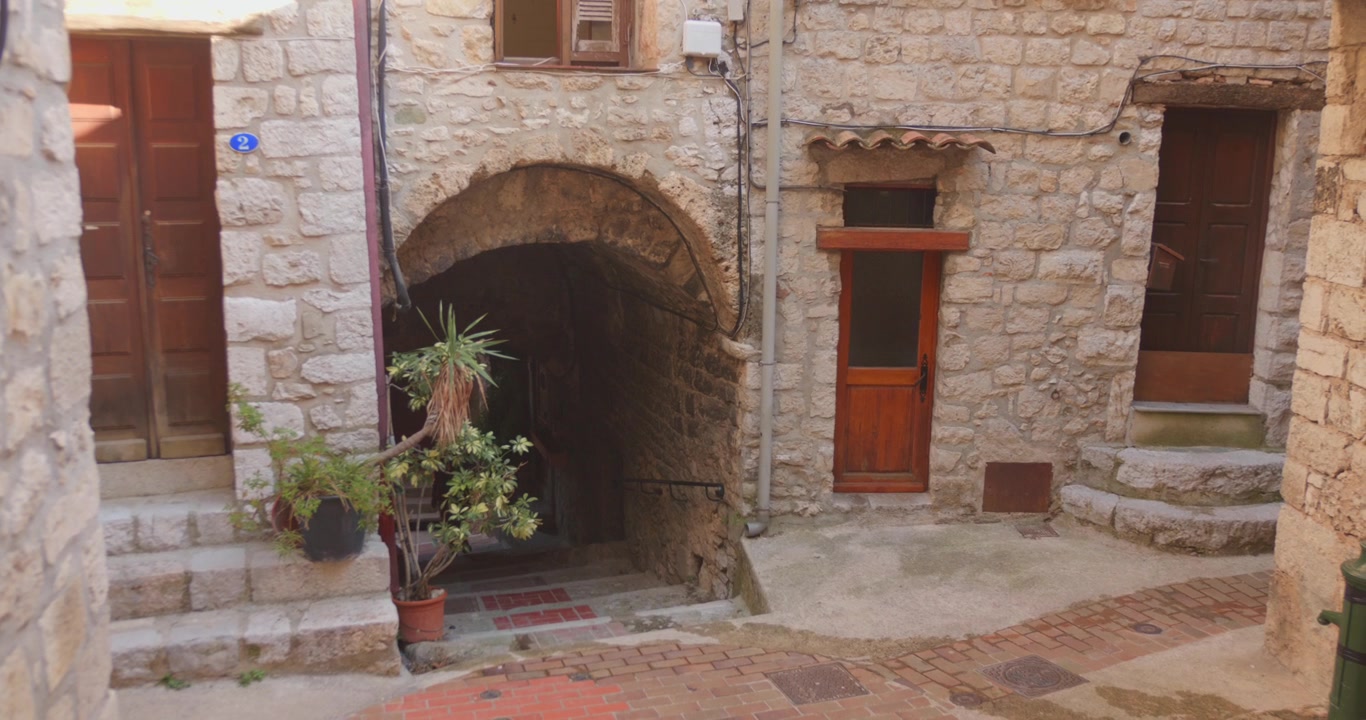 Tranquillo vicolo in pietra con scalinata ad arco, piante in vaso e porte in legno nel borgo medievale di Peille, nel sud della Francia
