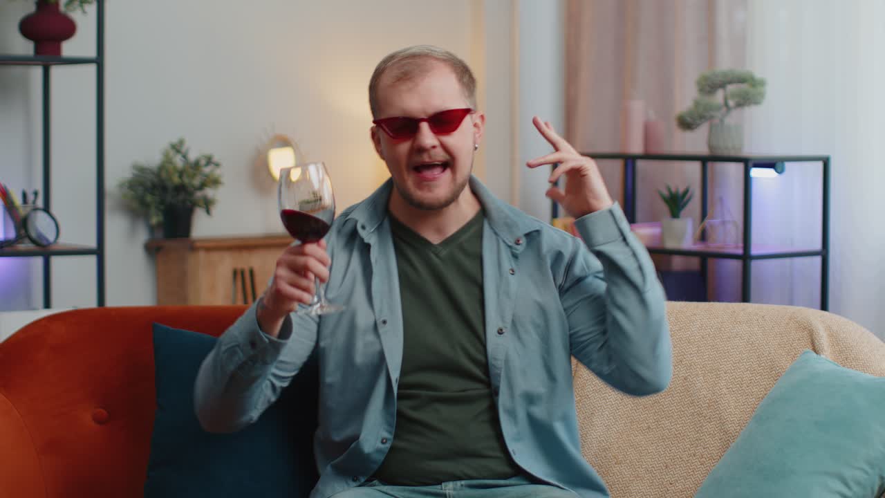 웃는 젊은 성인 남자가 은 와인 잔을 마시며 생일 기념일을 축하하고 집에서 복권에 승리합니다.