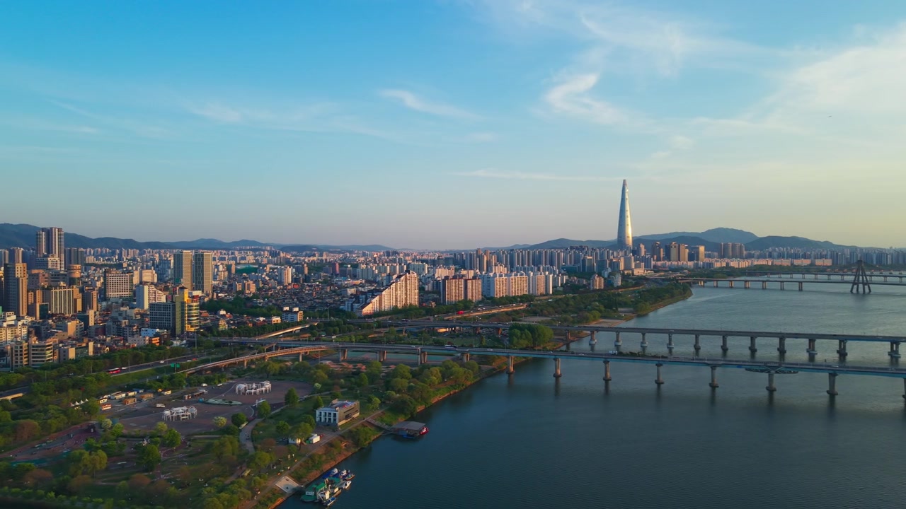 Una panoramica aerea al tramonto si estende dai ponti Gwangjin e Cheonho attraverso il fiume Han verso il Parco Gwangnaru Hangang, rivelando la Lotte Tower che risplende di luce dorata.