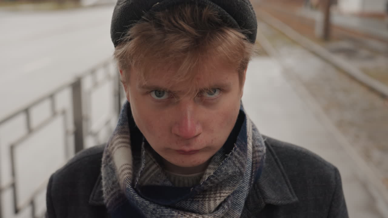 White creator head bowed on platform, damp pavement and passing cars, knit cap and layered scarf, intimate closeup revealing pale skin and focused eyes, tender cinematic mood with creative