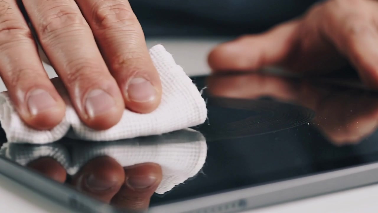 Asettaen taitetun kankaan, tummahihaiset miehen kädet pyyhkivät tabletin näyttöä valkoisella pöydällä, poistaen tahroja.