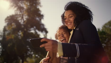 Lachende-Mutter-Hält-Smartphone-Und-Schaut-Sich-Lustige-Bilder-Im-Park-An.