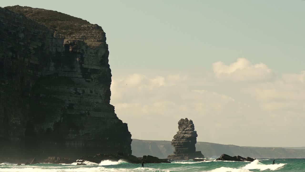 People paddle boarding on low waves next to a tall cliff on a sunny day