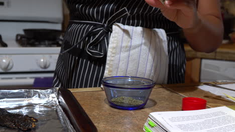 Woman-Measuring-Oregano-into-Small-Glass-Blue-Bowl,-Close-Up