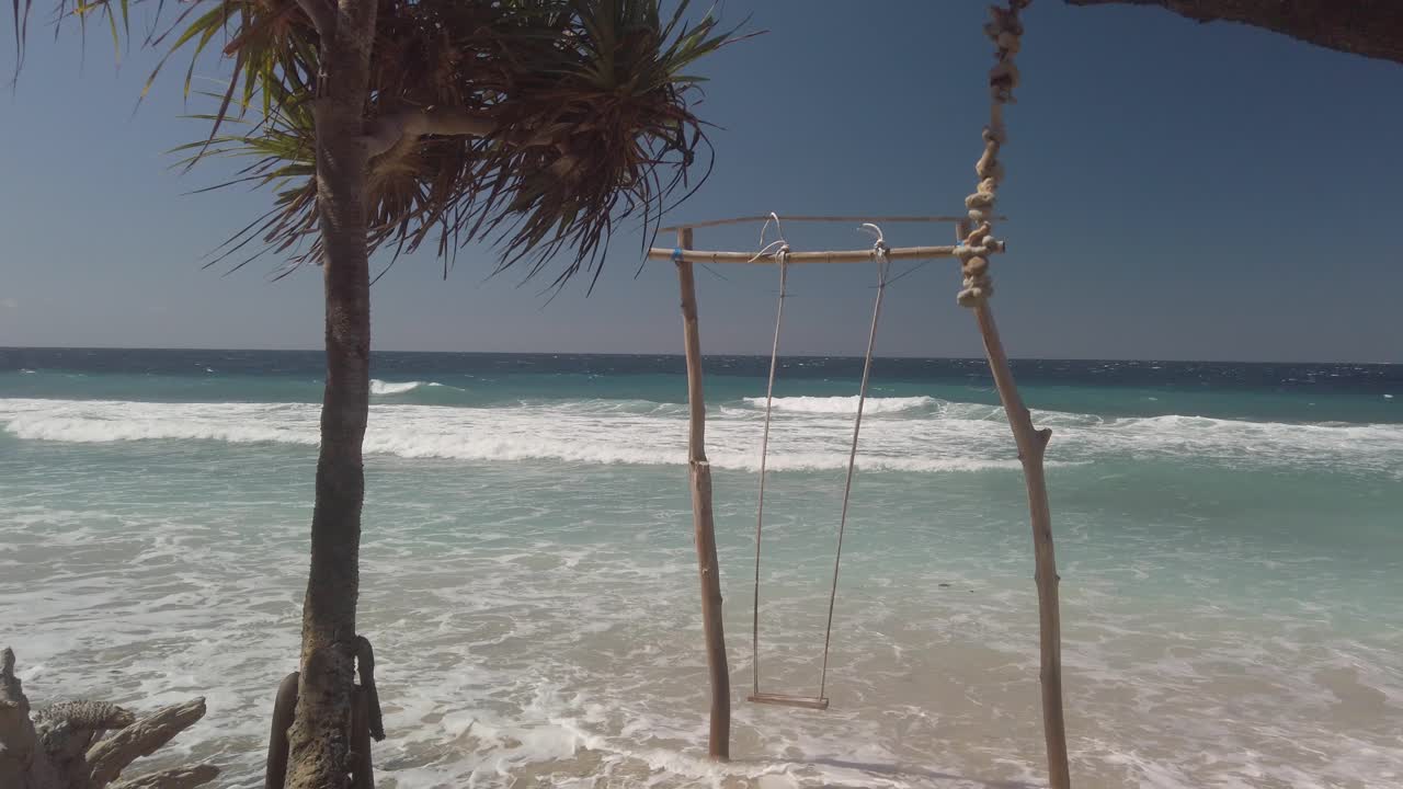 Balanço de praia em uma praia de areia com o azul do oceano ao fundo, ao lado de uma palmeira exótica. Filmagem manual de um dia ensolarado e ventoso em Bali, Indonésia.