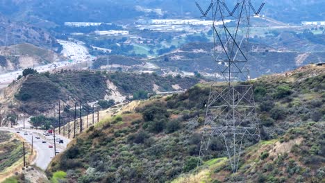 Aerial-view-rising-up-electricity-power-pylon-stretching-across-traffic-travelling-winding-valley-highway-in-the-distance