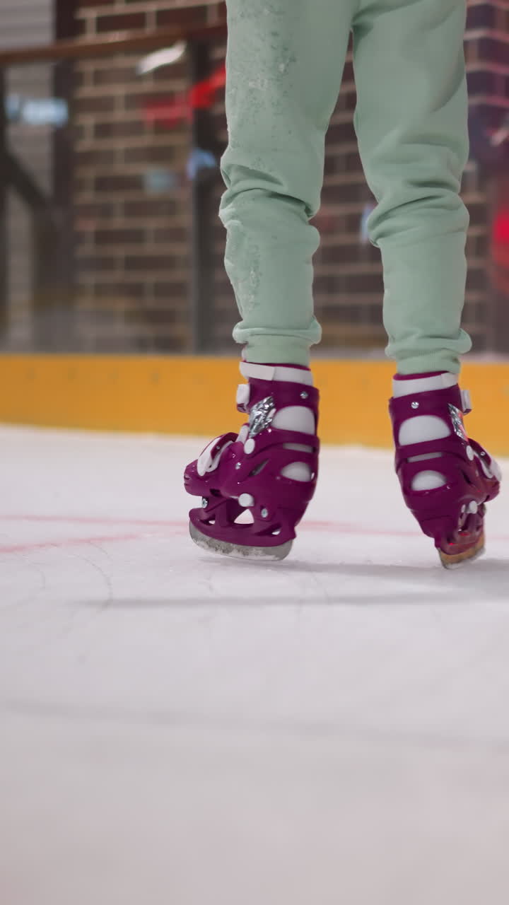 vista cercana de dos personas patinando en una pista de hielo, una con patines púrpuras y pantalones verdes menta, la otra con patines azules y pantalóns negros, contra un telón de fondo de luces rojas y una pared de ladrillo