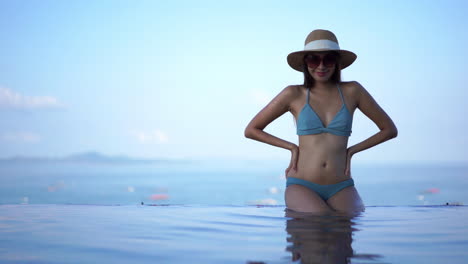 Young-fit-Asian-woman-stands-in-the-shallow-end-of-a-resort-swimming-pool-wearing-sunglasses,-a-sun-hat,-and-bikini-with-her-hands-on-her-hips-and-a-smile-on-her-face