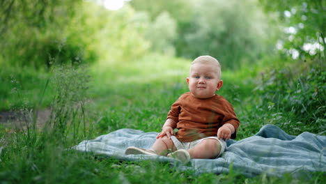 little blond baby boy with unhappy face sits on the plaid outdoors. toddler kid looks around at the green grass.