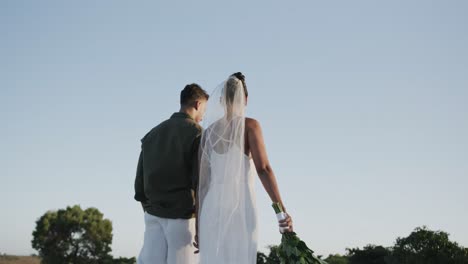 rear view of diverse bride and groom walking away holding hands at beach wedding, in slow motion