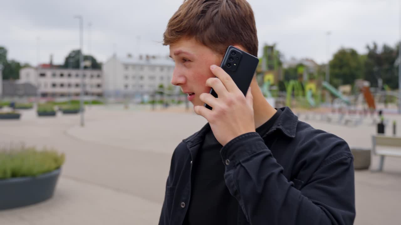 Young man talking on smartphone outdoors in city park square, serious phone call conversation, urban lifestyle, communication technology