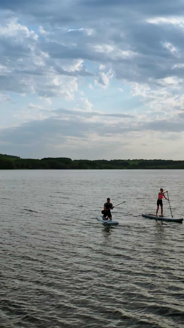 People spend time on water in the evening. Young man and woman sailing on river on boards with oar in summer at dusk. Orbital view. Vertical video