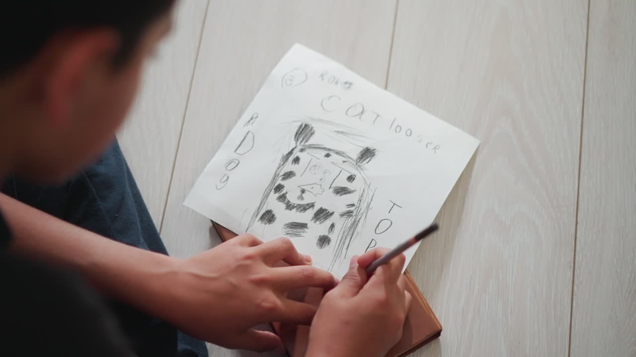 Primer plano de un niño dibujando un gato con un lápiz sobre un papel blanco apoyado en un libro marrón mientras está sentado en el suelo de madera, capturando la creatividad, la concentración, el juego y la imaginación de la infancia.