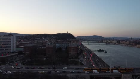 Vista-Aérea-Del-Tráfico-Al-Atardecer-Con-El-Río-Danubio-En-Budapest-Con-La-Ciudad-Y-Las-Montañas-Al-Fondo