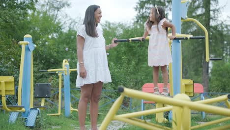 a pregnant mother and her young daughter enjoy playful time together at a playground in the park, surrounded by trees and greenery