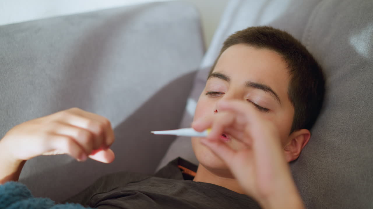 Close-up of sick boy collecting thermometer to check his temperature, concerned expression, covered with blanket, indoor setting with soft lighting, child health monitoring moment
