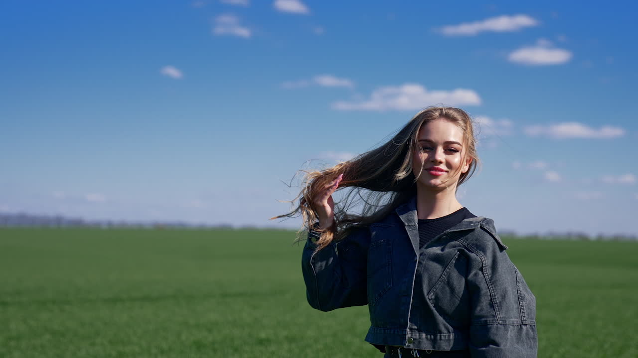 Attractive model with fluttering hair on field. Smiling young woman walking and looking on camera on green nature background. Slow motion.