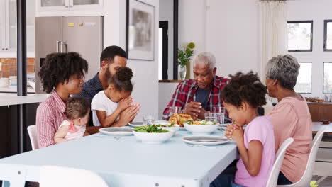 Familia-Multigeneracional-Orando-Antes-De-Comer-Alrededor-De-La-Mesa-En-Casa