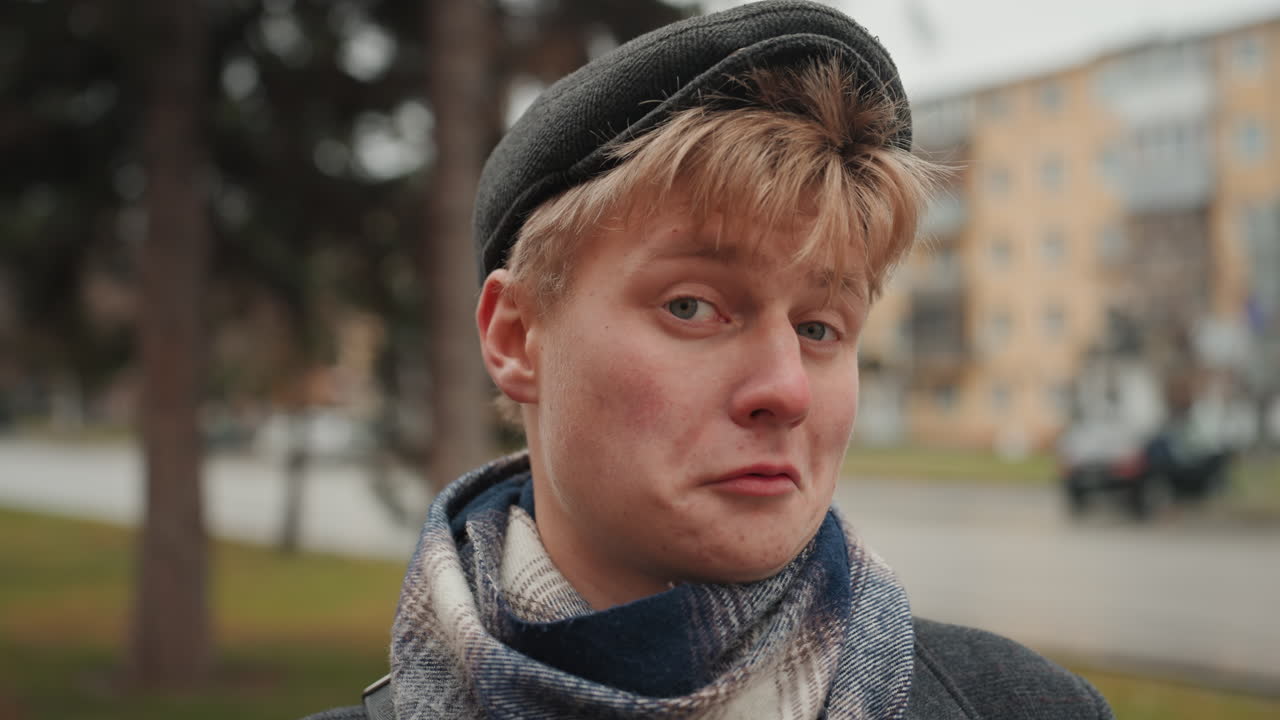Young man smelling red rose on street with trees and apartments, contemplative closeup portrait, scarf and beret styling, soft bokeh background, cinematic intimate mood, creator admiring bloom