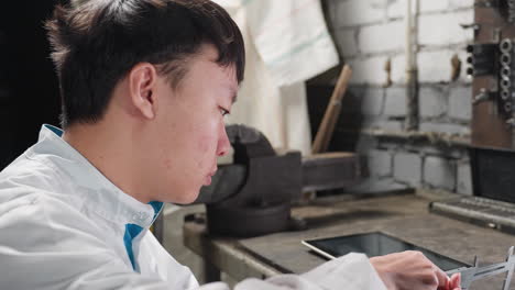 young lab technician working with veiner caliper adjusting measurements, tapping on tablet, returning to adjust caliper on boris placed on table, background includes mechanical tools