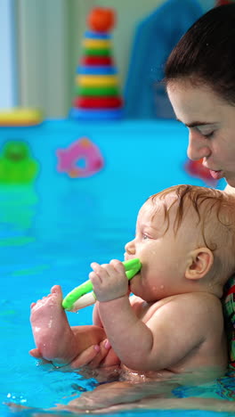 woman with a little baby boy in the swimming pool. adorable infant holds the toy in his mouth. side view. vertical video