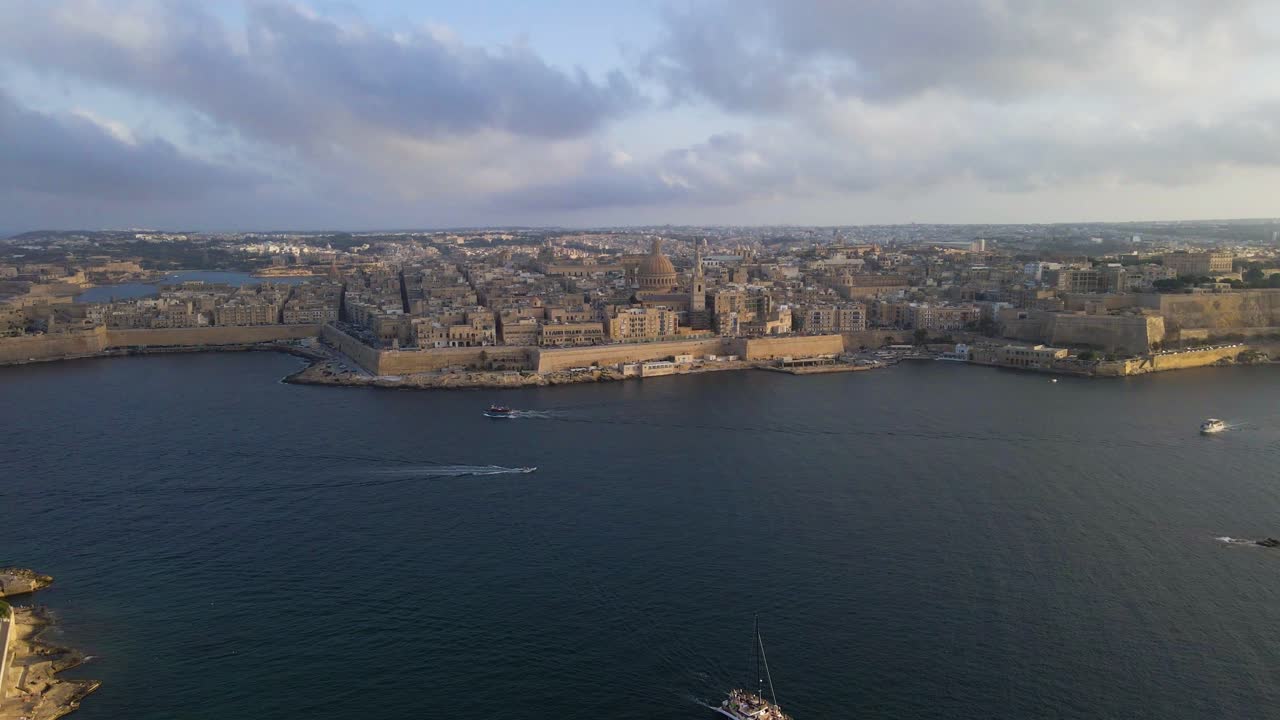Epic wide aerial drone footage of Valletta's complete fortified peninsula surrounded by deep blue Mediterranean waters, with Grand Harbour, historic bastions, and a lone sailboat under dramatic cloudy