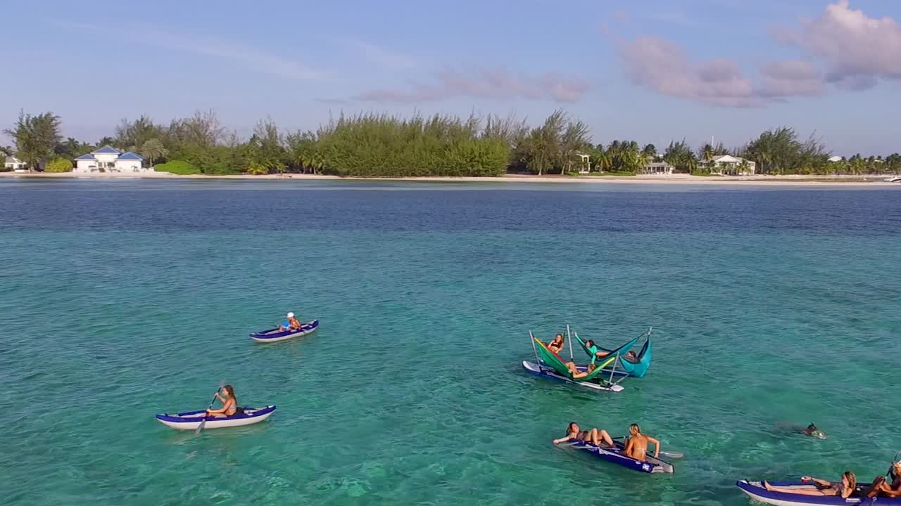 Drone flies over hammocks on wakeboards and kayakers paddling in clear turquoise water