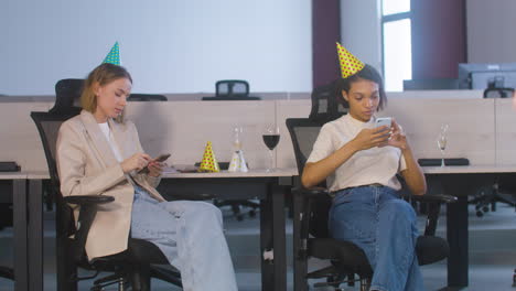 two female colleagues sitting in office chairs and using mobile phone during a party at the office