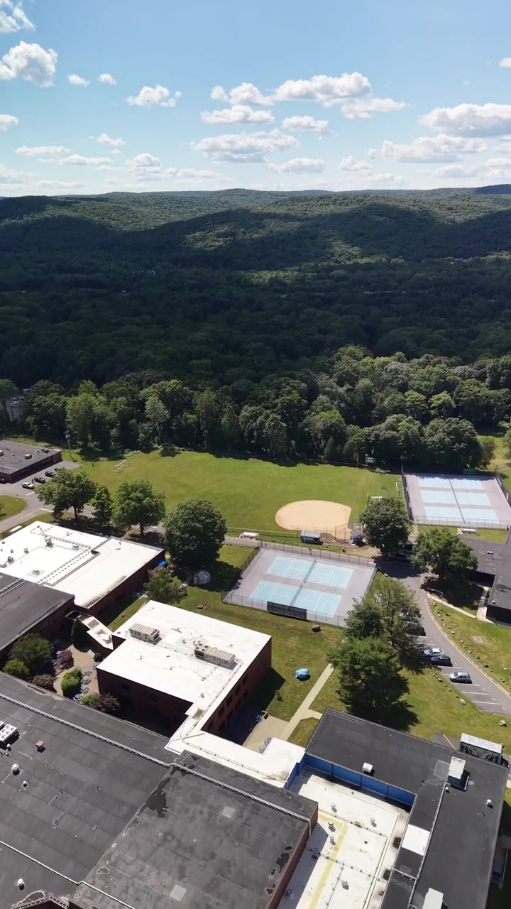 Aerial perspective showcasing a large educational building with a parking lot, alongside adjacent tennis courts and a baseball field, set against a backdrop of dense green forest and hills