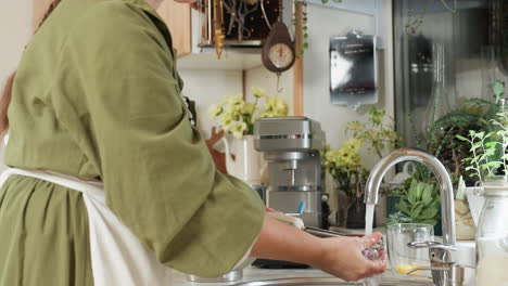 chef in green gown and white apron rinses electric mixer under running tap in bright modern kitchen surrounded by plants and utensils after cooking, ensuring cleanliness and proper kitchen hygiene