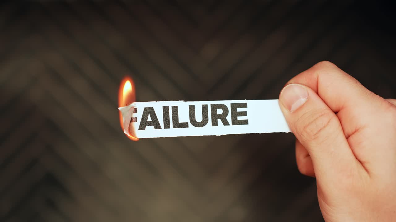 Close-up of a hand holding a paper strip labeled “FAILURE” as a flame burns the edge on a dark background—symbol of letting go, resilience, and mindset shift with copy space