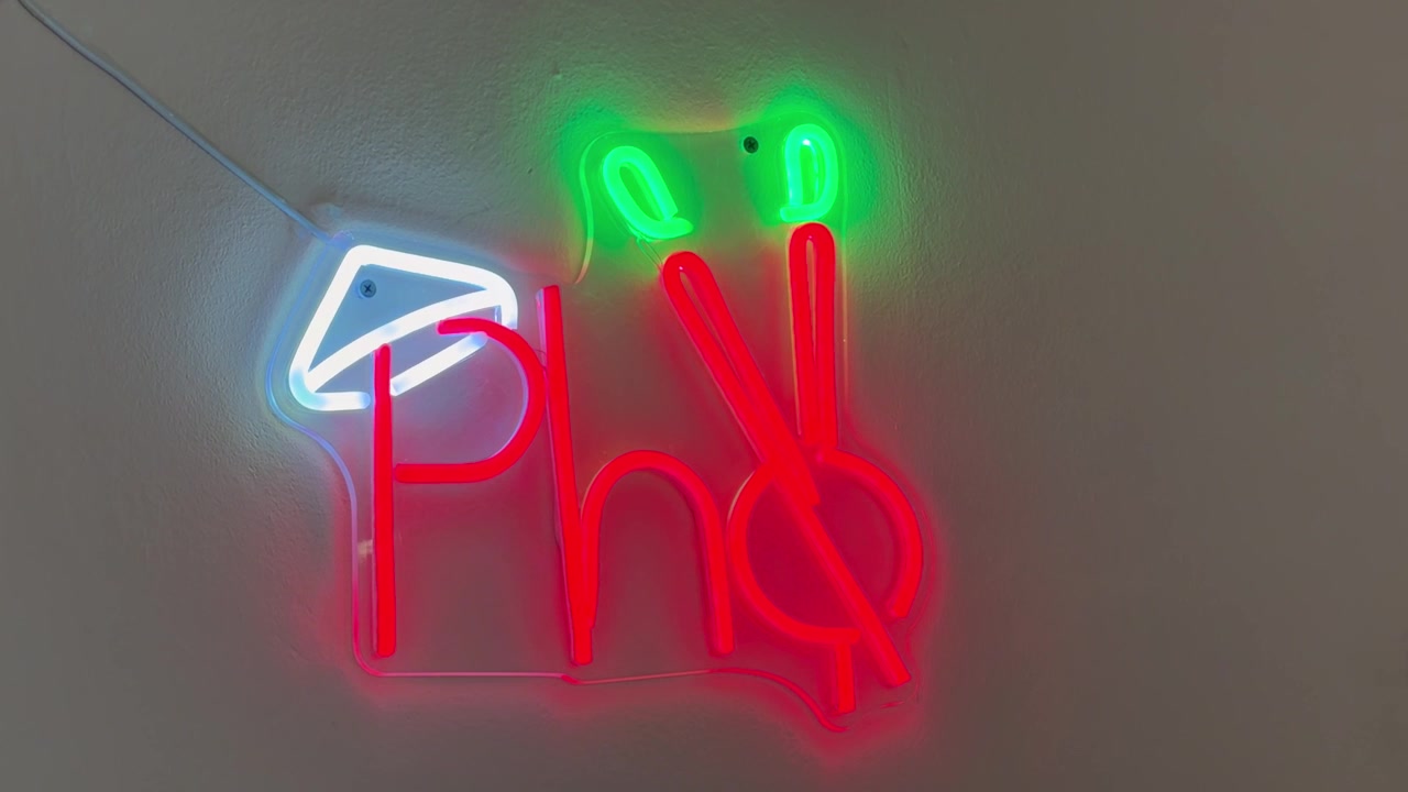質感のある壁に取り付けられた、明るい赤色のチューブで「Pho」と書かれた鮮やかなネオンサインのクローズアップショット。上部には緑色のアクセント照明があり、左側には白い幾何学的な形が配されている。