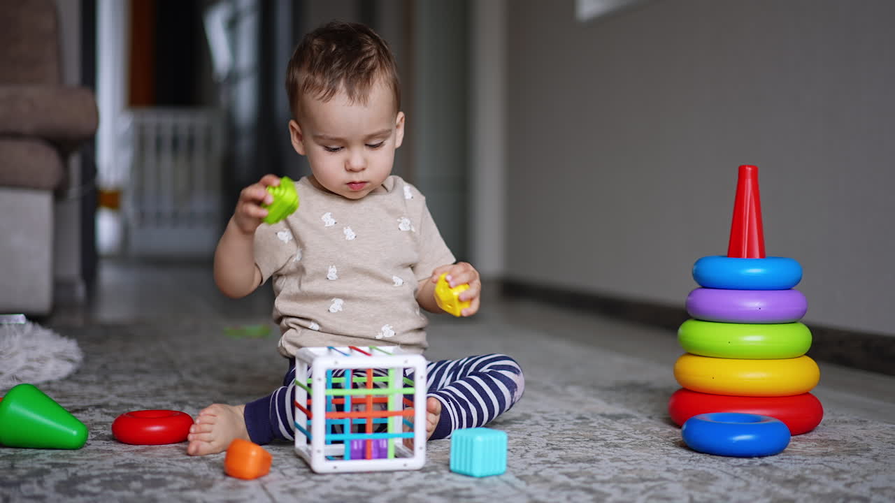 Serious baby boy plays with the toys sitting on the carpet. Toddler playing peacefully at home.