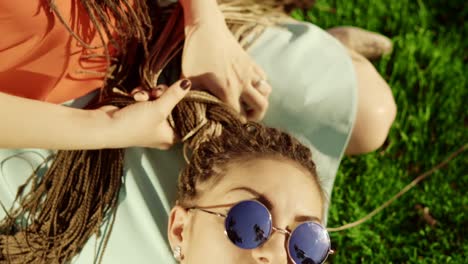 young hipster girls relaxing on the grass in park. one girl is weaving braids for another. two hipster women lying on the grass