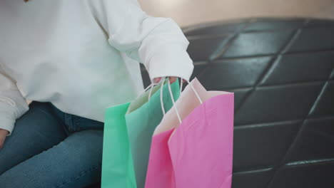 vista parcial de una mujer con un traje blanco colocando bolsas de compras rosas y menta en un asiento de cuero en un centro comercial se sienta cerca de ella mientras coloca su mano en su regazo con un fondo borroso