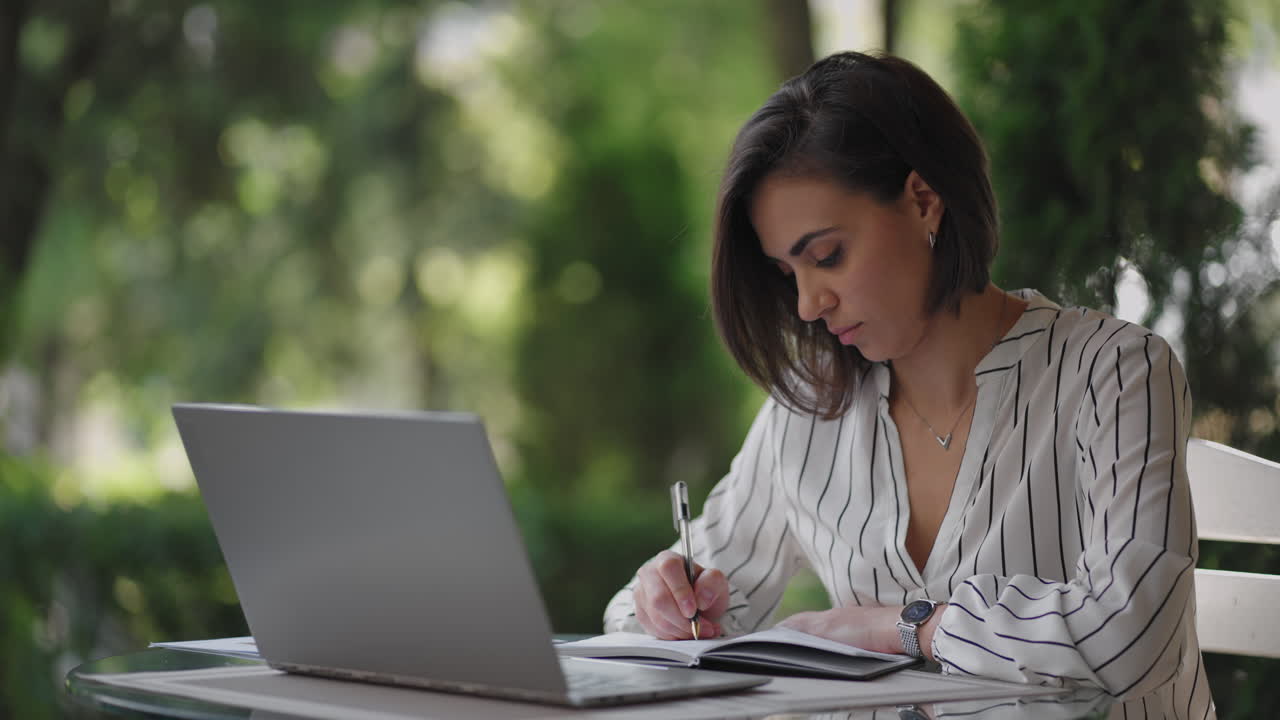 mujer de negocios morena de etnia hispana trabaja remotamente mientras está sentada en un café de verano en un día soleado con una computadora portátil y escribe con un bolígrafo y un cuaderno