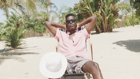 smiling african american man sitting on deckchair wearing sunglasses on sunny beach, slow motion