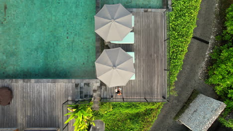 Young-Asian-Woman-In-Dress-Walking-Along-Swimming-Pool-at-Tropical-Island-Hotel-in-Bali-Indonesia,-Aerial-Top-Down-View