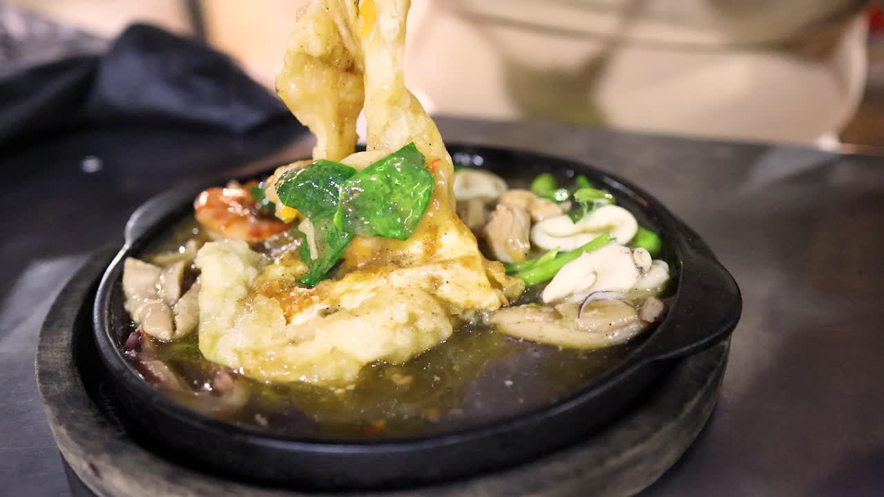 Hands stir sizzling fried noodles with vegetables and meat on hot plate, vibrant night lighting