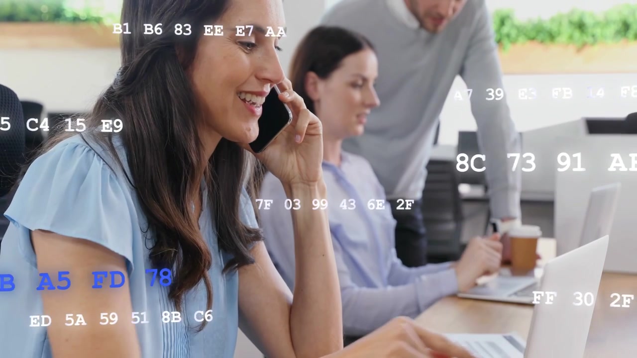 Woman receiving call, alternating typing and speaking at shared desk, code overlay aiding tech work