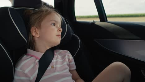 niña caucásica de 6 años sentada en el asiento de seguridad y mirando por la ventana del coche durante un viaje en coche.