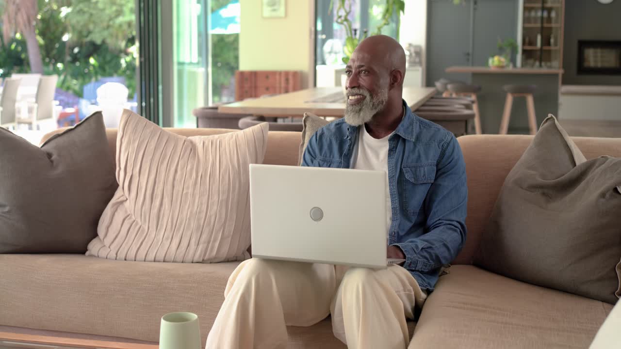 Senior African American man opening silver laptop, typing on sofa, checking email, glancing outside