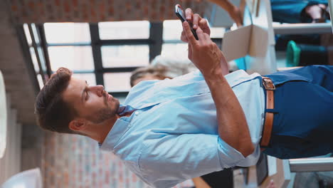 Video-Vertical-De-Un-Hombre-De-Negocios-Parado-En-Una-Oficina-Ocupada-Enviando-Mensajes-Por-Teléfono-Móvil