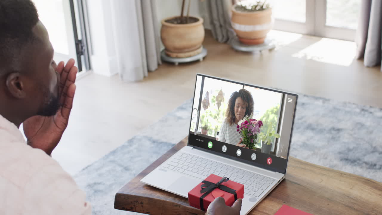 African american man holding red gift using laptop with african american woman and flowers on screen