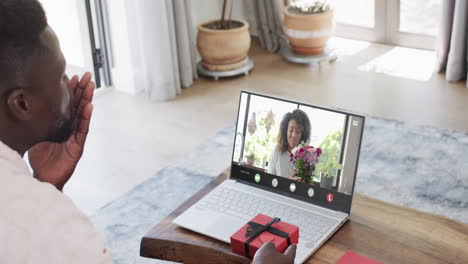 Hombre-Afroamericano-Sosteniendo-Un-Regalo-Rojo-Usando-Una-Computadora-Portátil-Con-Una-Mujer-Afroamericana-Y-Flores-En-La-Pantalla