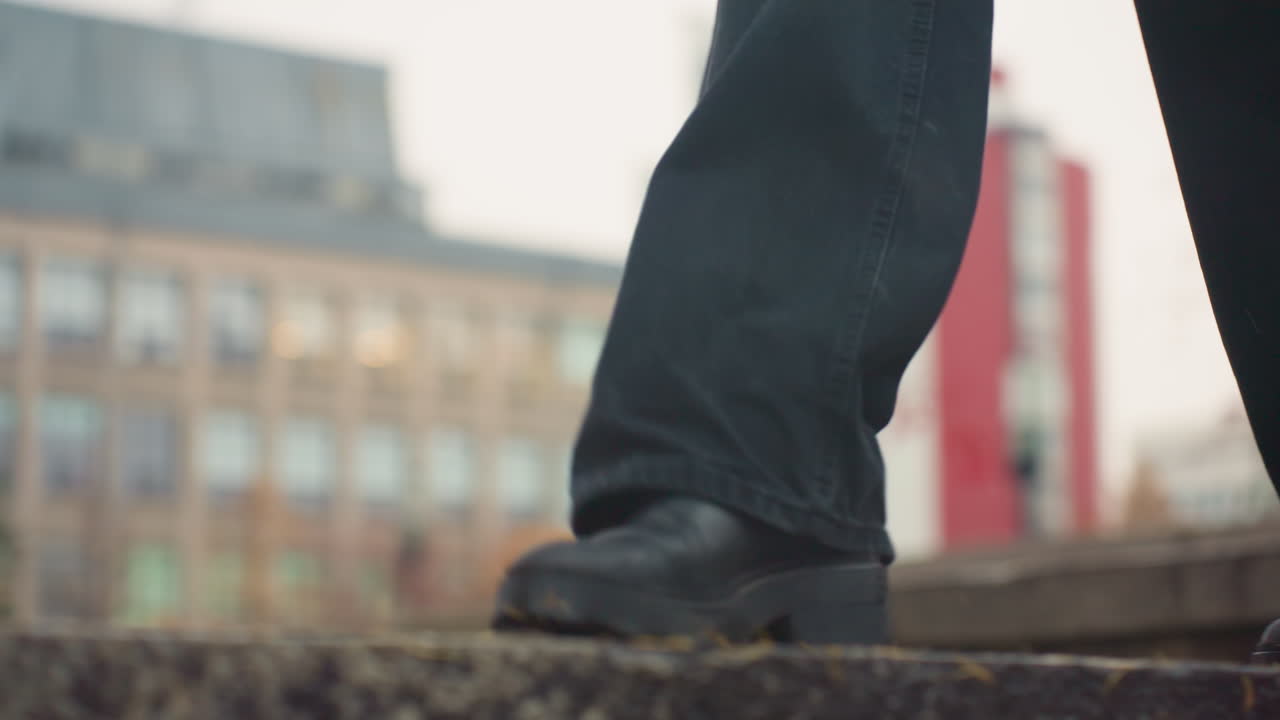Close-up view of person in black trousers and black boots carefully walking on wide wet stone path during rainy autumn day, maintaining balance amidst the rain and leaves