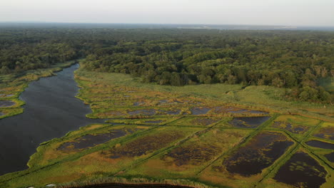 Grúa-Descendente-Disparó-Sobre-Las-Aguas-En-El-Puerto-Deportivo-De-East-Islip---Parque-Que-Se-Centra-En-El-área-Del-Pantano-Con-Un-Canal-De-Chanel-Más-Ancho-A-La-Izquierda-Al-Atardecer,-Volando-Sobre-Algunos-Barcos-Atracados-En-El-Muelle-De-Madera-Debajo