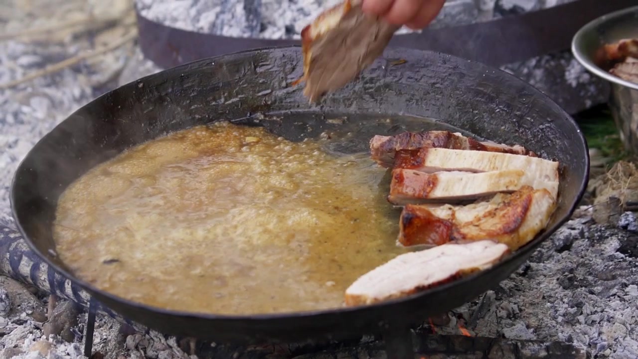 Zbliżenie na plastry wieprzowiny rumieniące się na żeliwnej patelni nad otwartym ogniem, w plenerze, przy naturalnym świetle, z widoczną dłonią, w rustykalnym stylu gotowania.